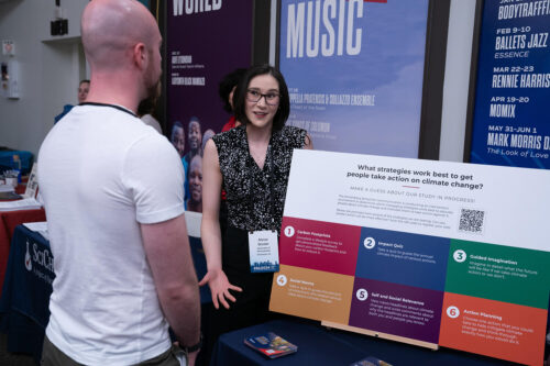 Alyssa Sinclair stands next to a table with a poster labeled "What strategies work best to get people take action on climate change?" and talks to a person in front of table.